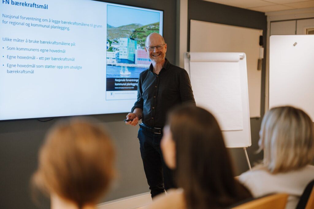 En mann som holder en presentasjonskontroller og smiler til publikum. Bak mannen henger det en stor skjerm som viser en presentasjon.