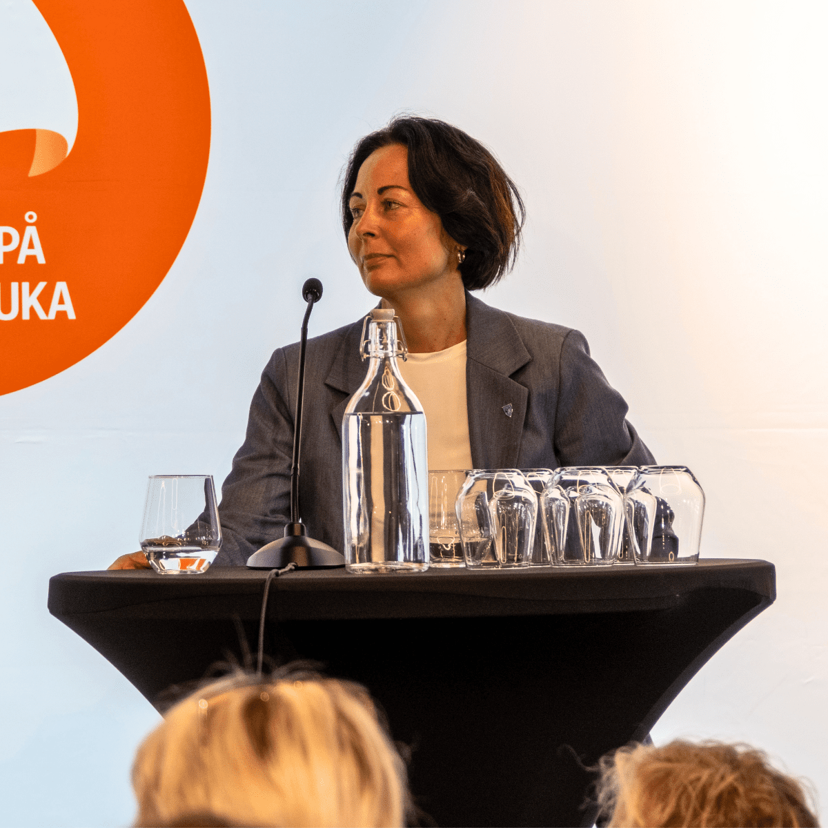 Kristine Dahl står bak et podium og ser til høyre for seg. En glassflaske med vann og mange glass står på podiumet.